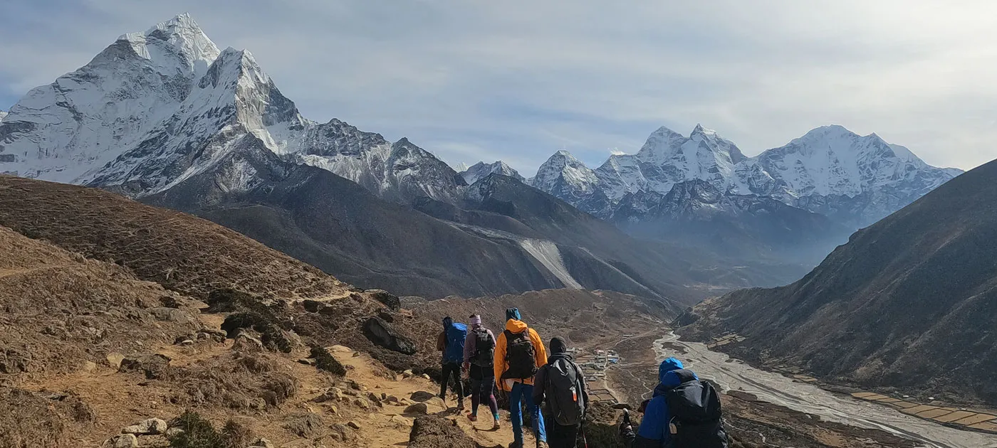 Group of hikers with backpacks walking down a mountain trail toward snow-capped Himalayan peaks including Ama Dablam