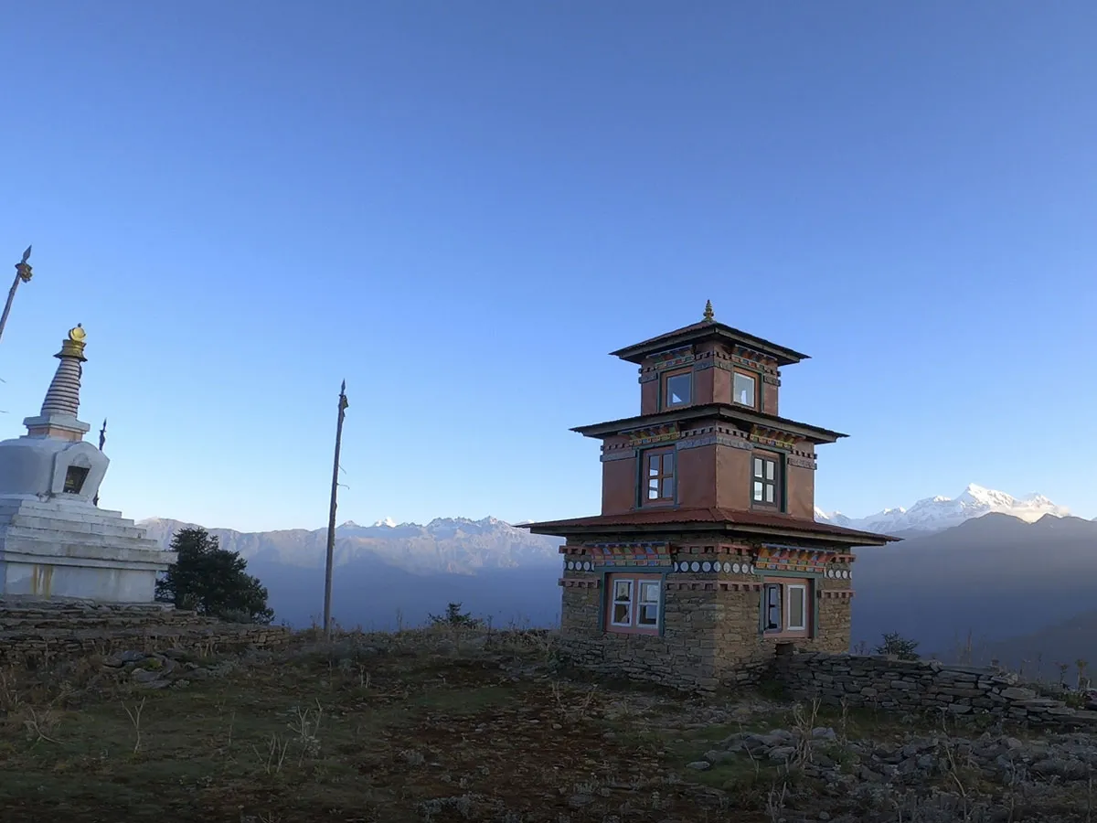 A stone Buddhist monastery building and a white chorten perched on a hilltop overlooking distant mountain ranges during clear daylight