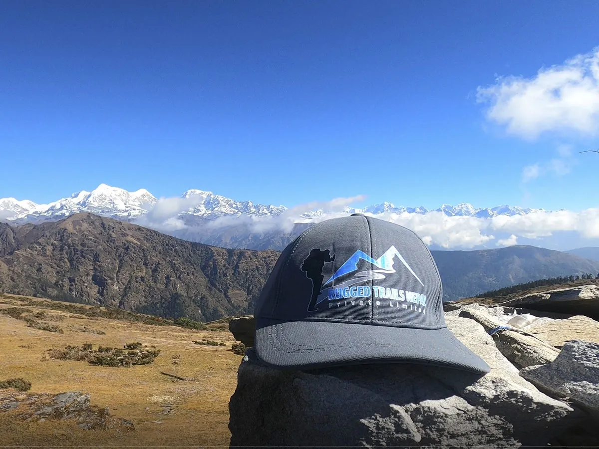 A grey trekking cap resting on a rock ledge overlooking a snow-covered Himalayan mountain range and open ridge pasture under a clear sky