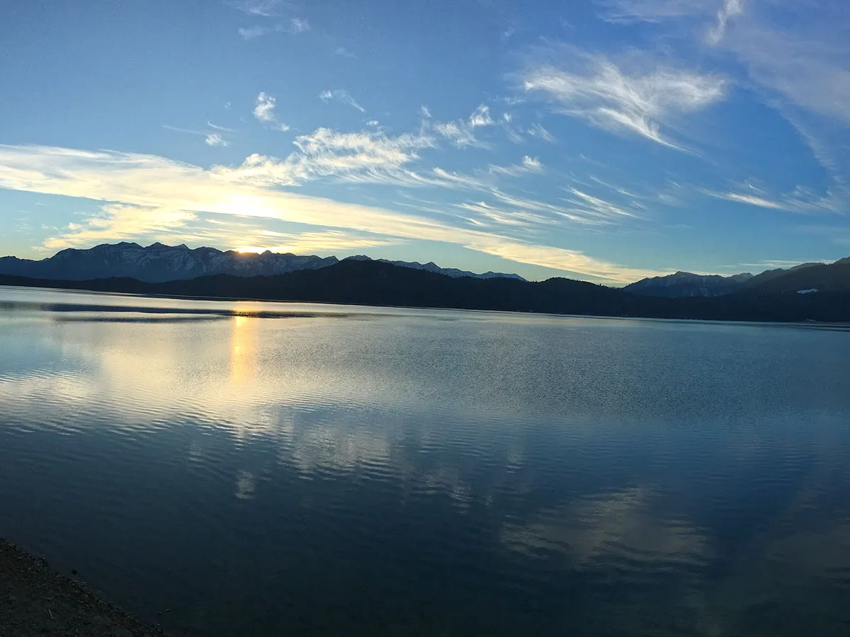 Panoramic wide shot of sunset glowing behind mountain silhouettes across the vast surface of Rara Lake under a light-filled sky