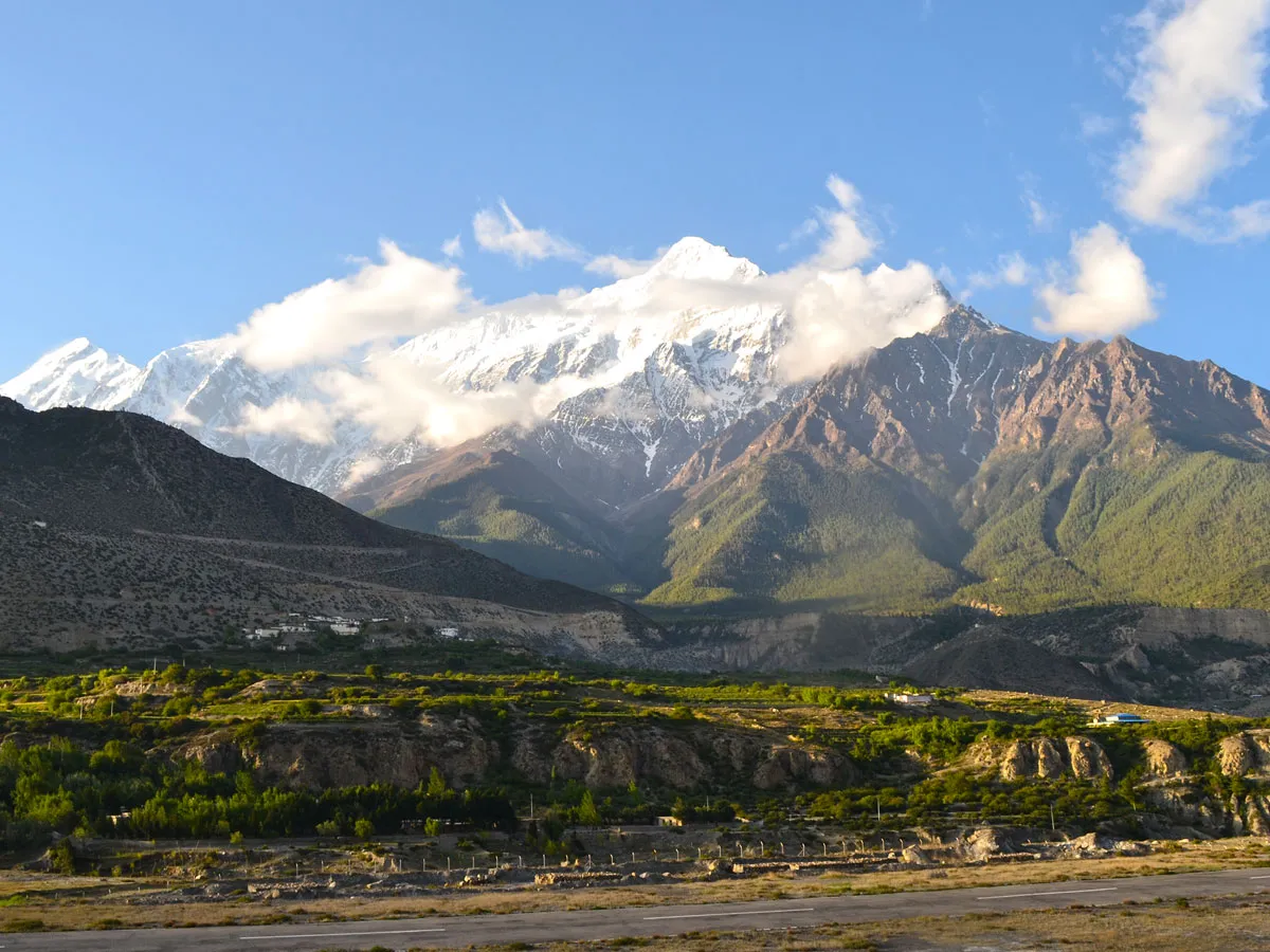 A majestic snow-covered Himalayan peak rising above green terraced fields, mountain slopes, and a paved valley road under a partly cloudy blue sky