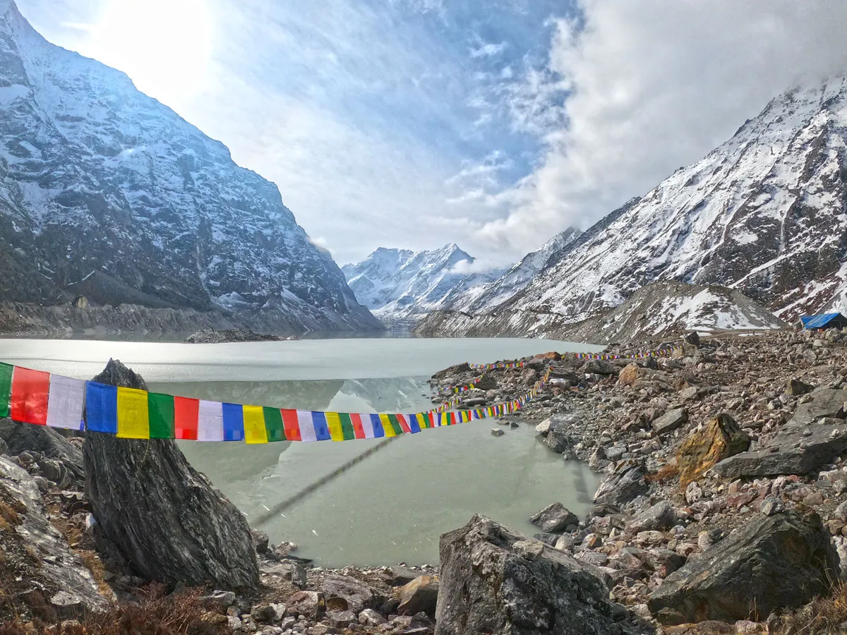 Vibrant Tibetan prayer flags strung across a rocky shore overlooking the icy, turquoise surface of Tsho Rolpa glacial lake surrounded by steep mountain walls