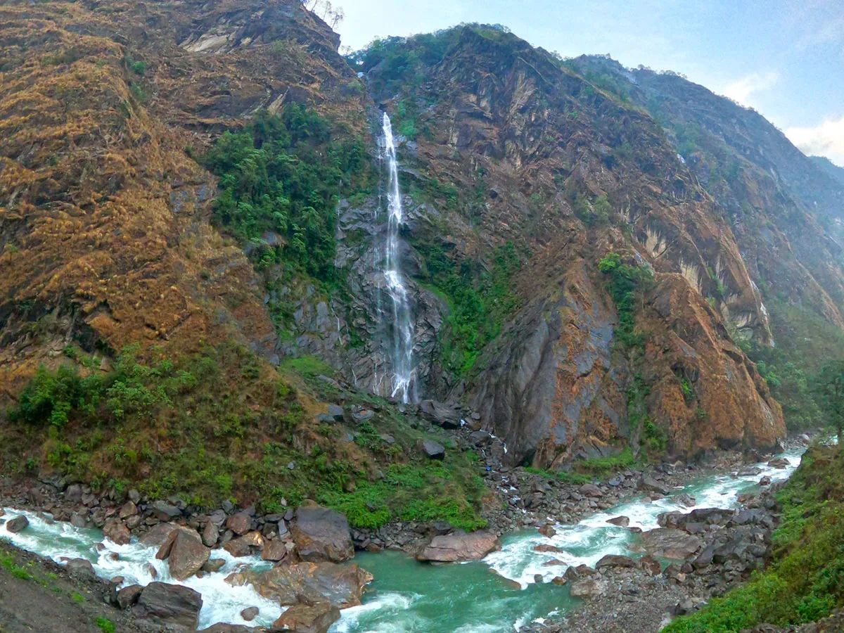 A tall narrow waterfall cascading down a sheer brown rock face into a rushing turquoise river in a mountain gorge