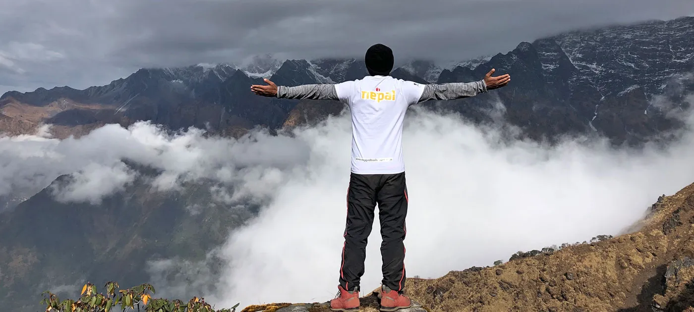 A trekking guide wearing a white shirt with arms stretched wide standing on a high mountain peak looking out over clouds and mountain ranges