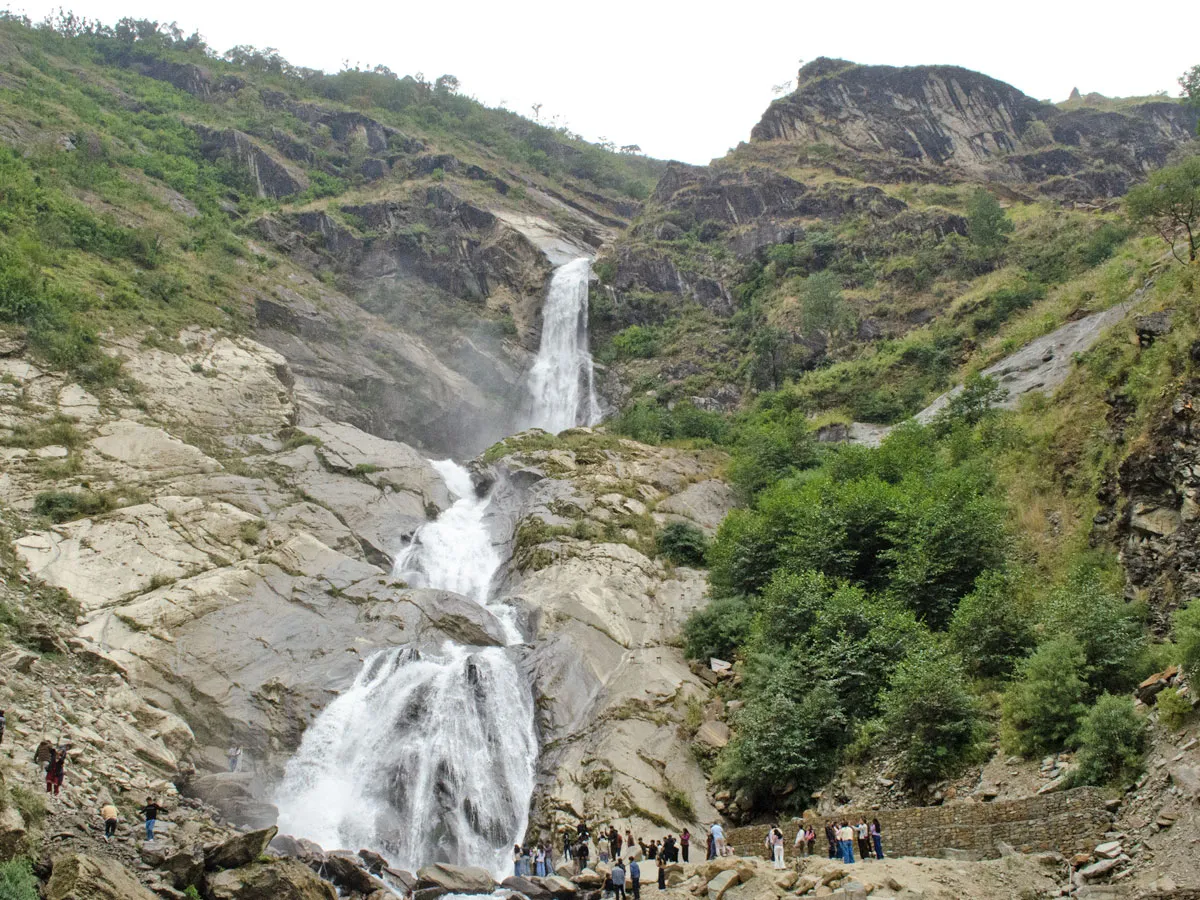 Powerful multi-tiered waterfall cascading down steep rocky cliffs surrounded by lush green mountain vegetation, with trekkers gathered at the base