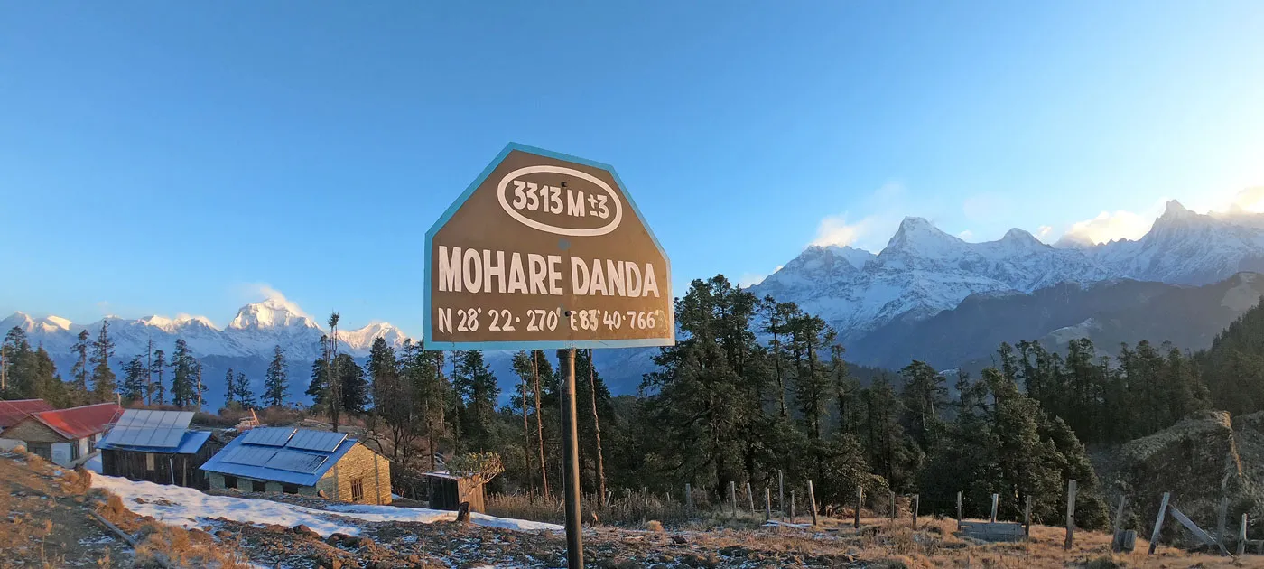 A wooden Mohare Danda elevation marker at 3,313 meters with solar-powered eco-lodges and snow-covered Himalayan peaks
