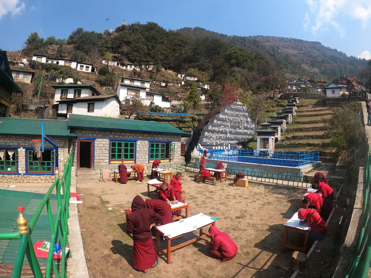 Young Buddhist monks in red robes sitting at wooden desks in an open-air monastery courtyard, studying and writing, with traditional Sherpa village houses and terraced hillsides in the background