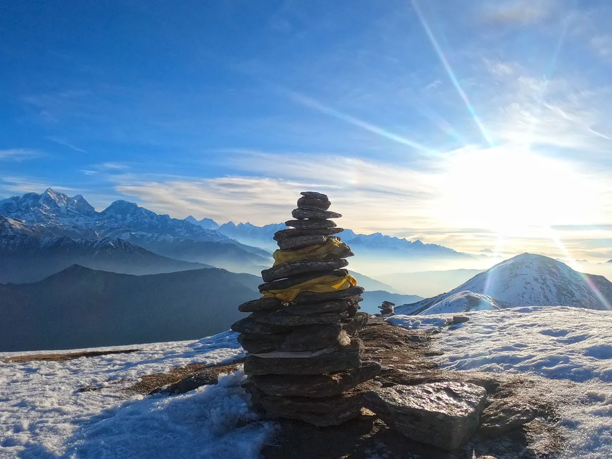 A traditional stacked stone cairn draped with yellow cloth on a snowy ridge, silhouetted against a bright sunburst and a panorama of distant mountain peaks