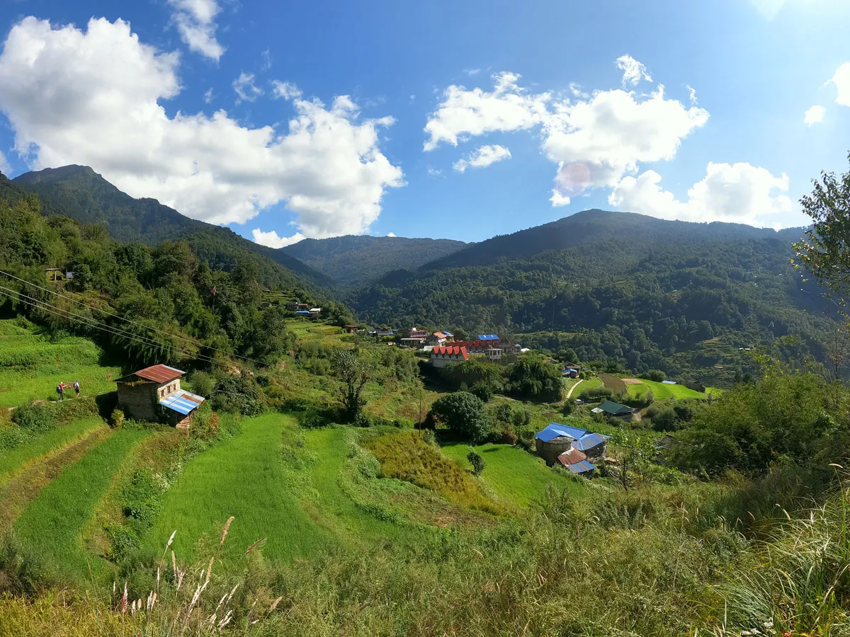 Panoramic view of a lush green traditional mountain village in Nepal with terraced agricultural fields and blue-roofed houses under a sunny sky.