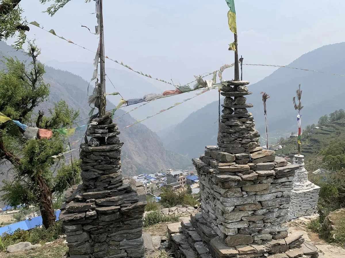 Traditional stone chortens and Buddhist prayer flags standing on a high ridge overlooking a mountain valley village.