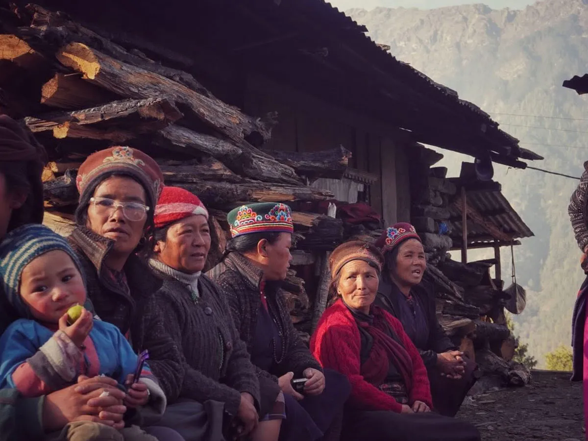 Local Tamang women wearing traditional embroidered headwear sitting outside a wooden village house with a young child.