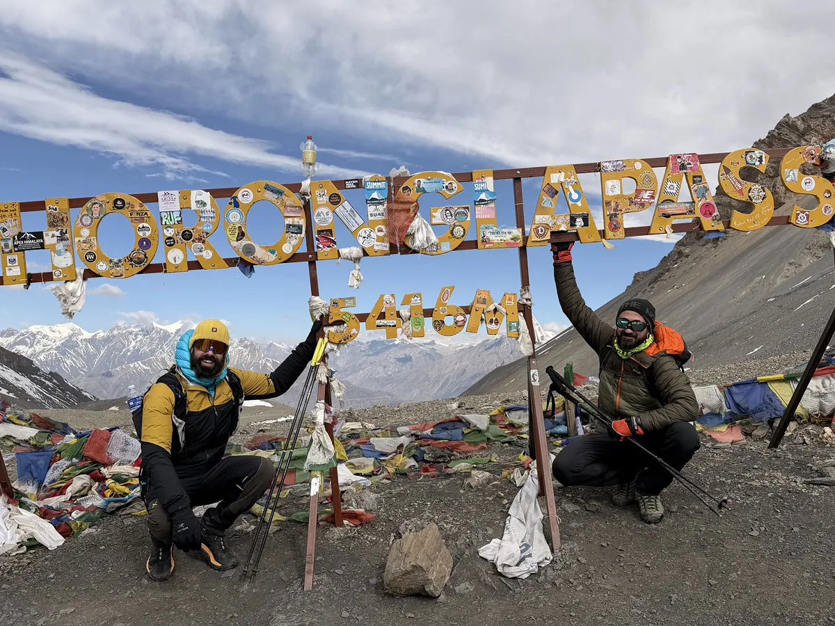 Two trekkers posing happily under the Thorong La Pass sign (5416m) decorated with stickers and prayer flags