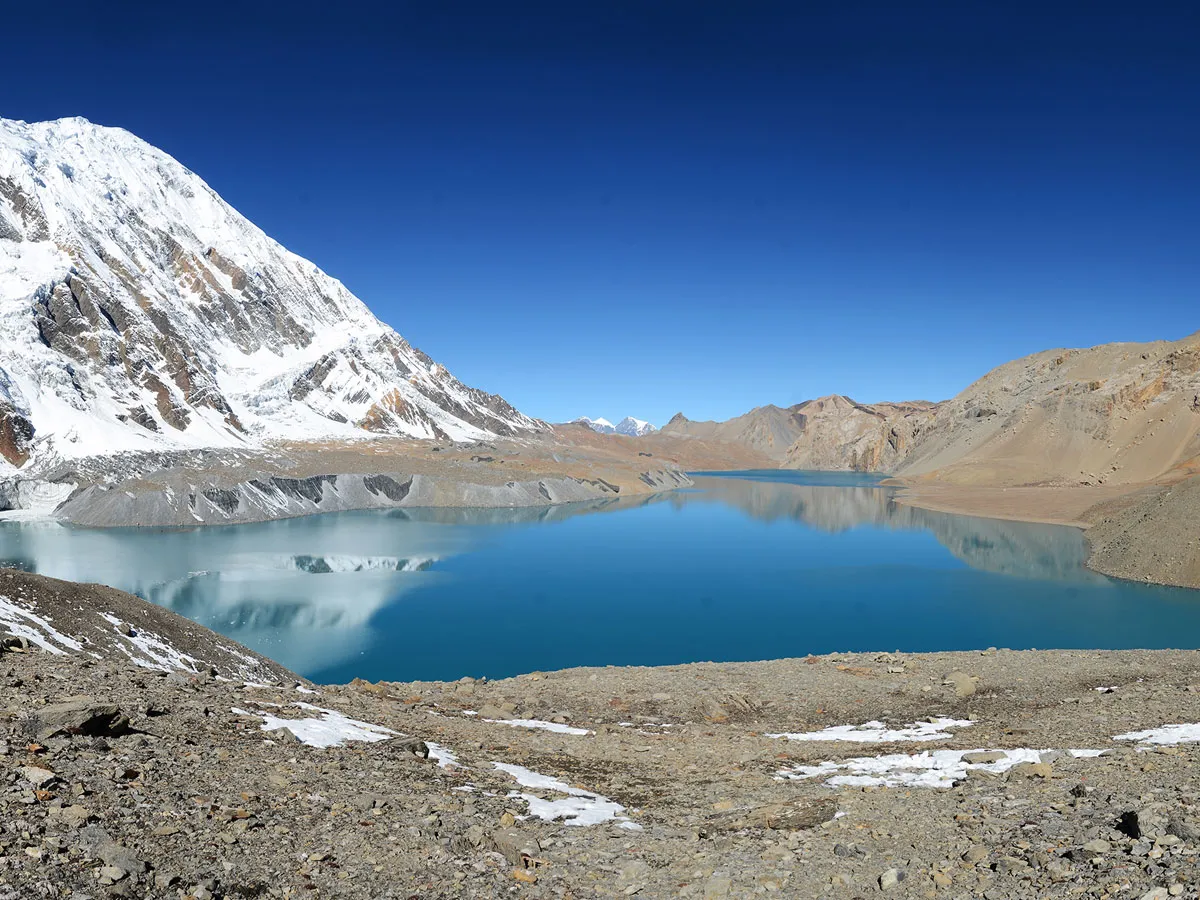 Turquoise waters of Tilicho Lake surrounded by high rocky terrain and snow-capped peaks under a clear blue sky in Manang, Nepal.