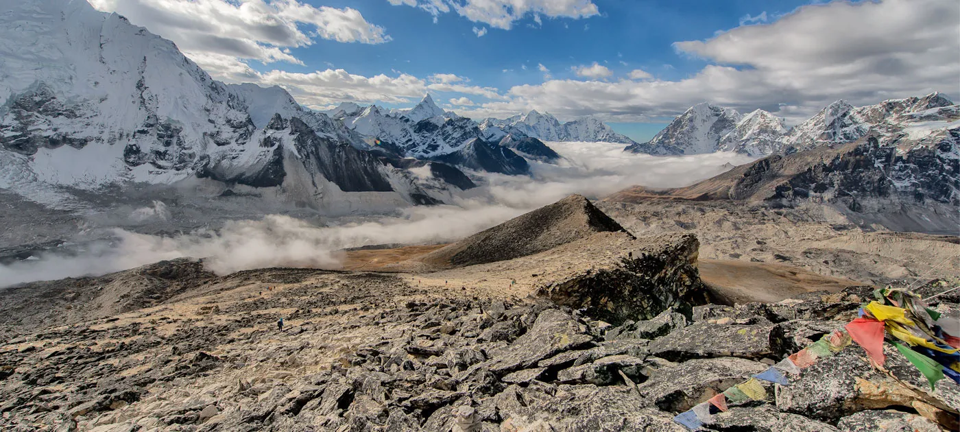 A high-altitude, rocky Himalayan landscape with colorful prayer flags in the foreground and clouds rolling through valleys below snow-capped peaks.