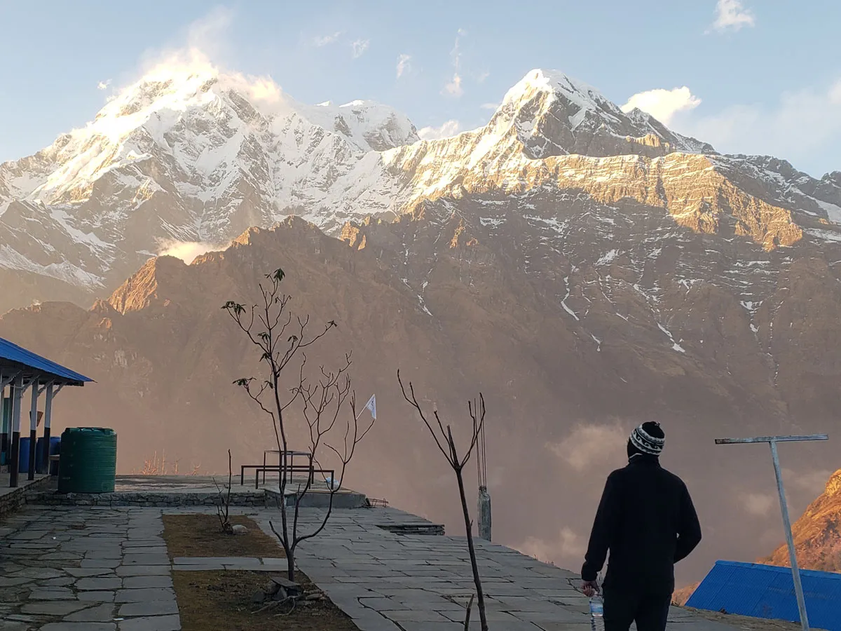 A trekker standing on a stone patio at a mountain lodge, looking out at golden sunlight washing over snow-capped mountain peaks.