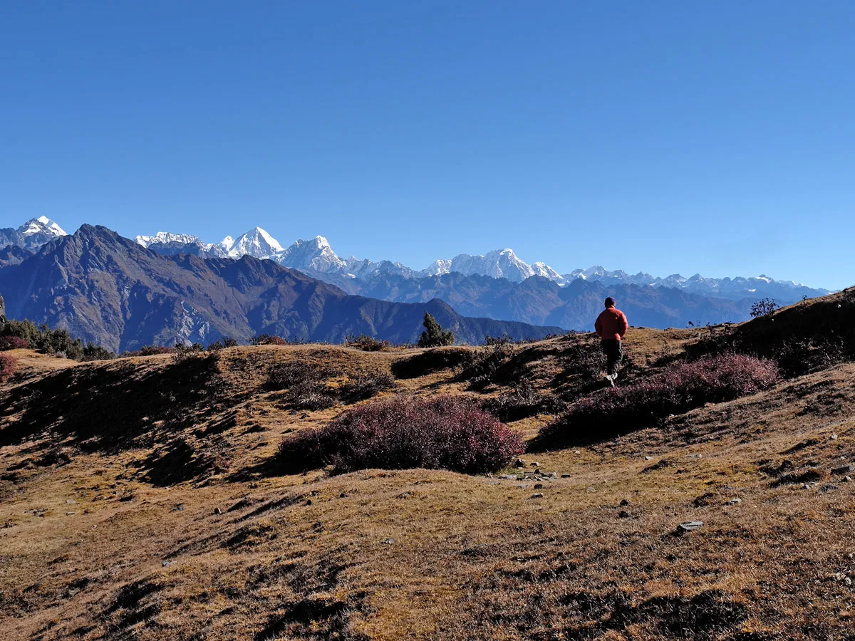 A trekker wearing an orange jacket walking along a ridge trail in the Helambu region with the Langtang mountain range in the background.