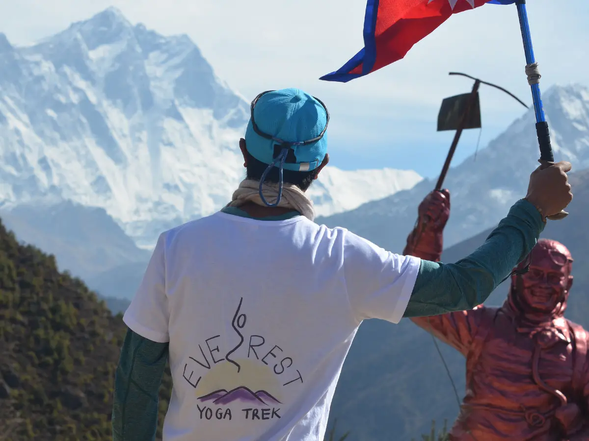 Trekker wearing Everest Yoga Trek shirt holding Nepal flag with Mount Everest in background.