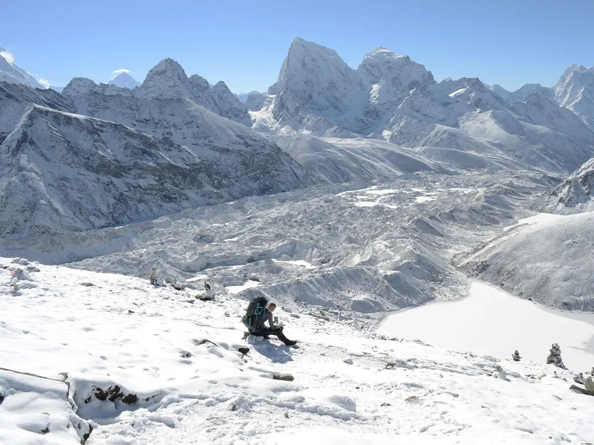 A trekker sitting on a snowy slope looking down at a vast glacier, frozen glacial lake, and surrounding snow-capped mountains in Nepal.