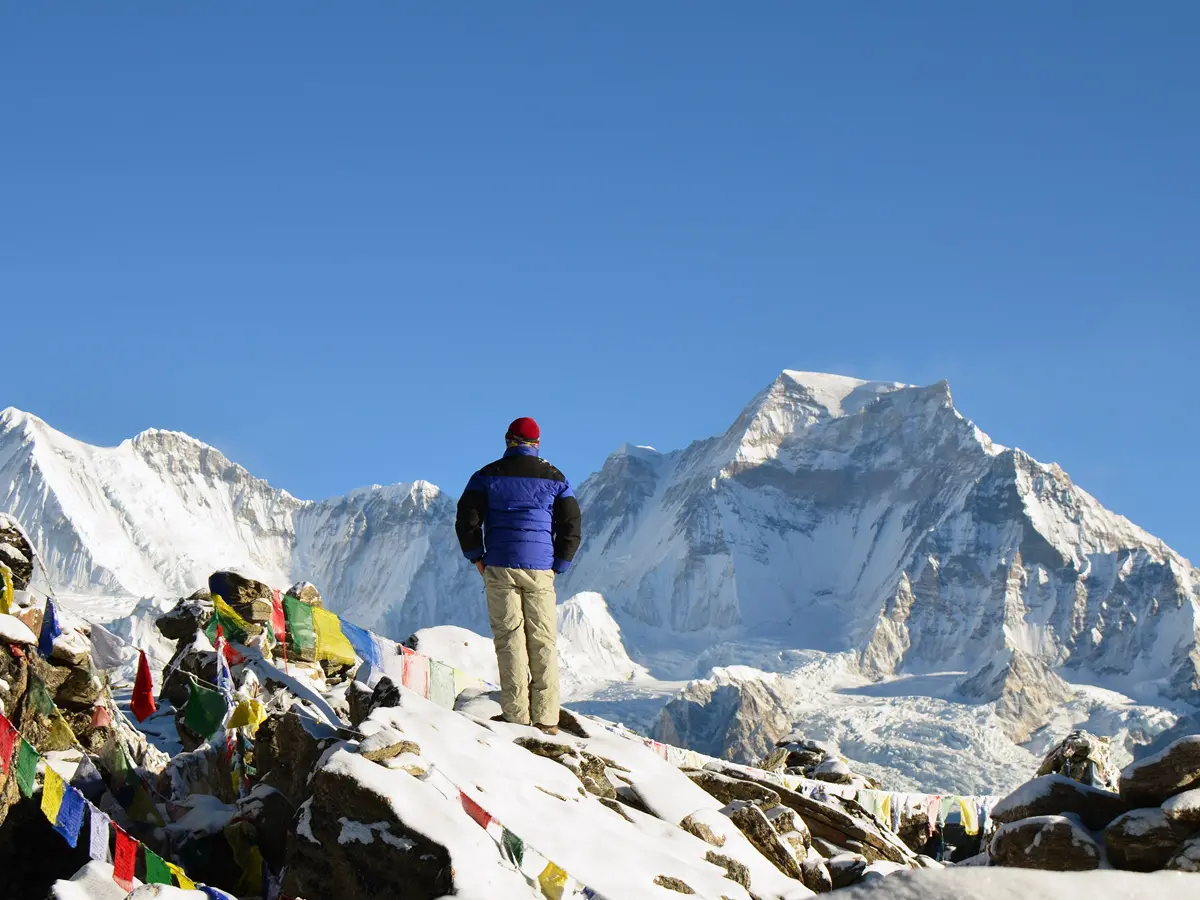 A lone trekker in a blue jacket and red beanie standing on a snowy ridge covered in prayer flags, gazing at massive Himalayan peaks