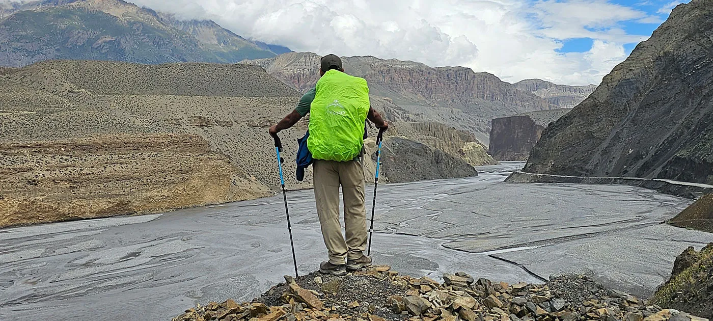 Rear view of a trekker with trekking poles wearing a bright green rain cover over his backpack, looking out over a river valley.