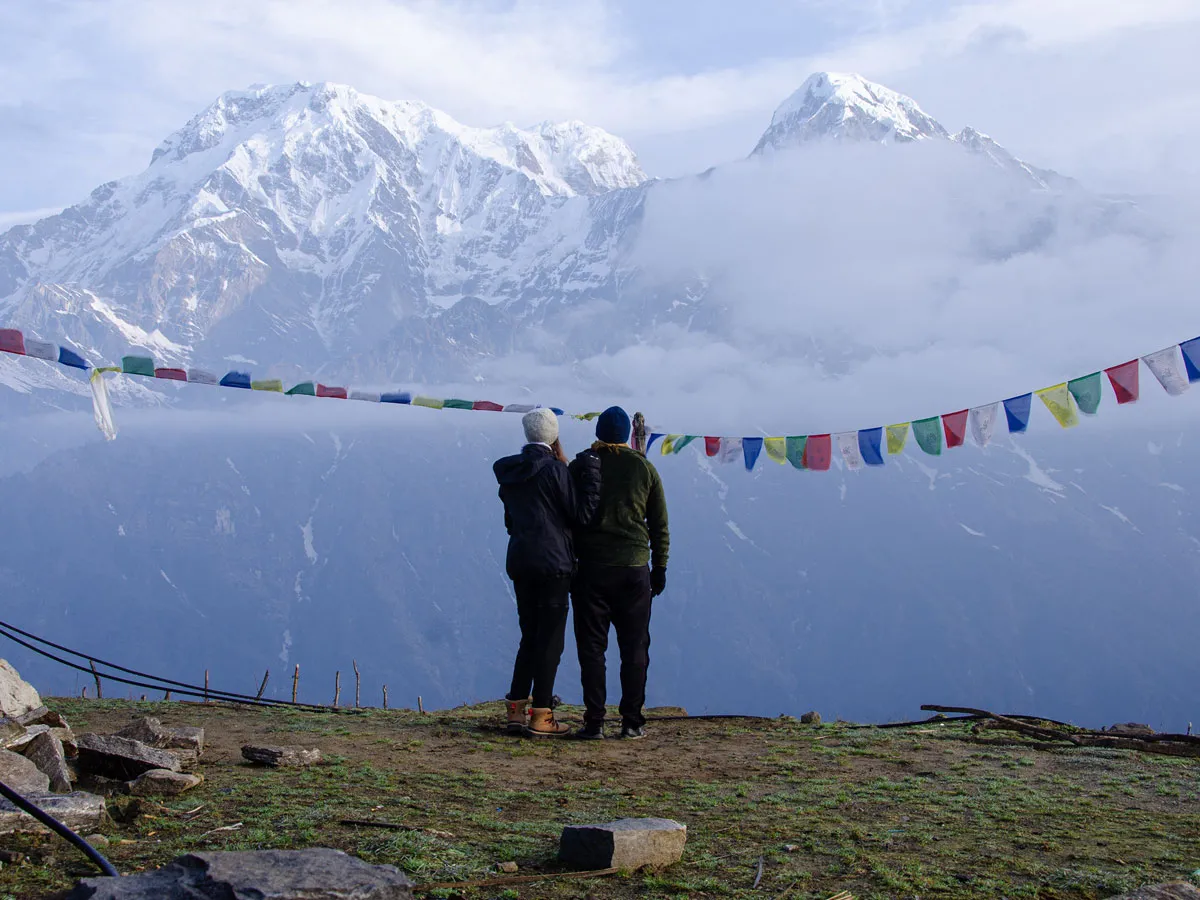 Two trekkers standing together behind colorful prayer flags, gazing at the misty Annapurna mountains on the Mardi Himal Trek.