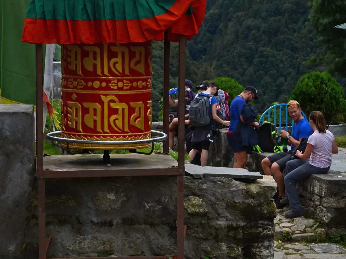 Group of trekkers resting on a stone trail next to a large red Buddhist prayer wheel.