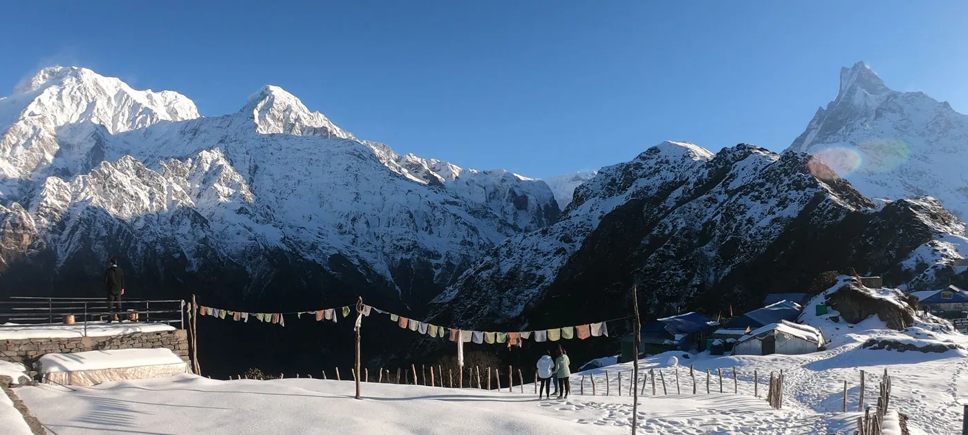 Panoramic view of a snow-covered Himalayan village with prayer flags and Mt. Machhapuchhre during winter trekking in Nepal.