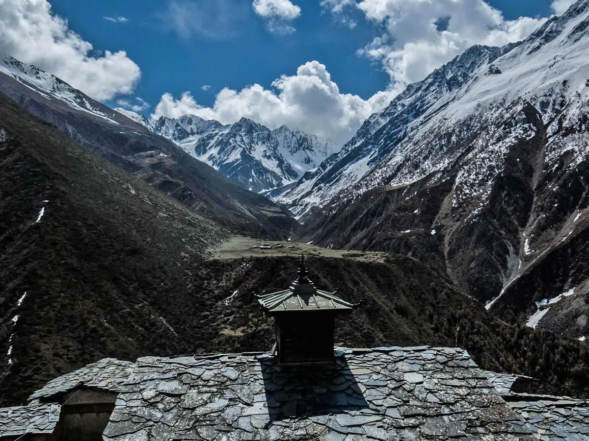 Slate tile roof with a central stupa spire overlooking a vast Himalayan valley and snow-covered mountain peaks under a blue sky