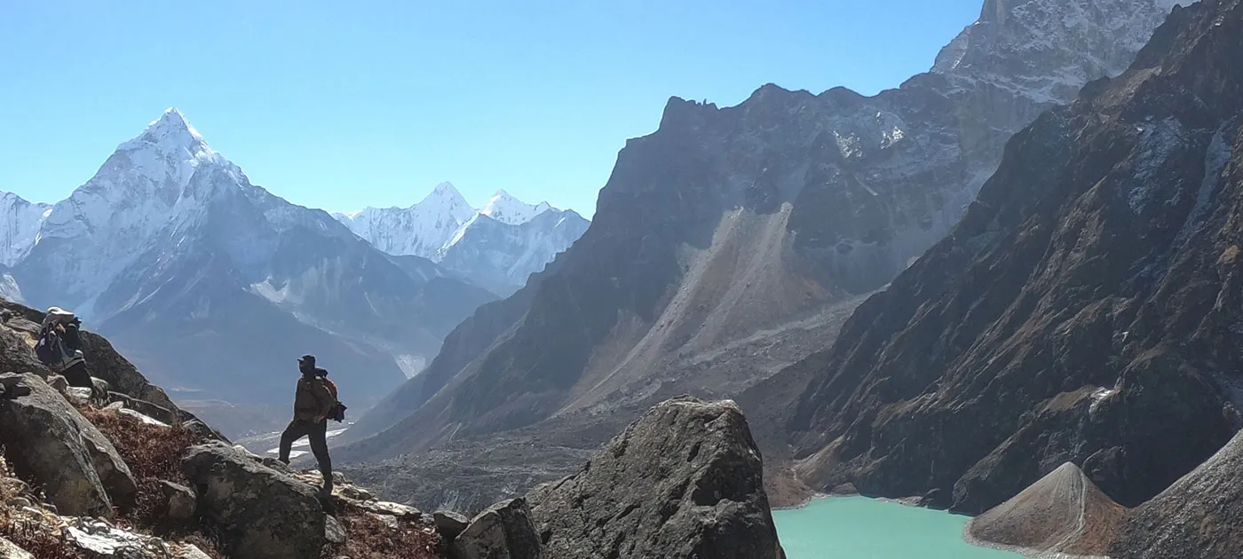 A trekker standing on a high rocky ridge looking down at a turquoise mountain lake and surrounding Himalayan peaks