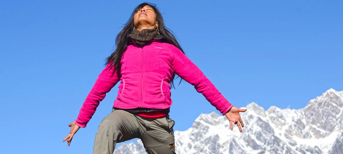 A woman wearing a bright pink jacket standing outdoors with arms outstretched and eyes closed in a peaceful pose in front of snowy mountain peaks