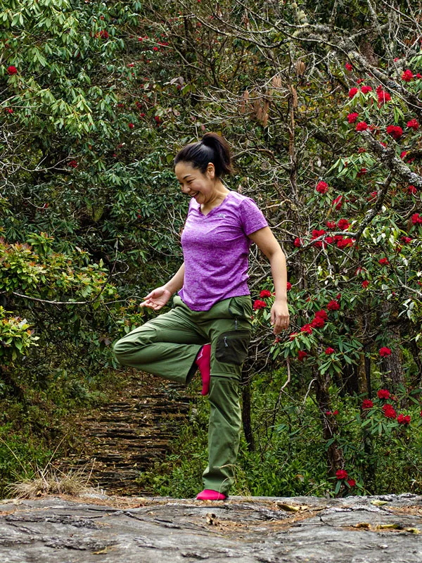 A cheerful female hiker posing on a tree-lined stone trail surrounded by blooming red rhododendron flowers in the Nepalese hills