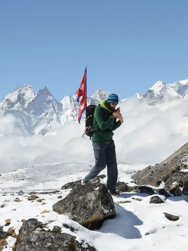 A hiker with a Nepal flag attached to their backpack navigating a rocky, snow-covered ridge high above the clouds