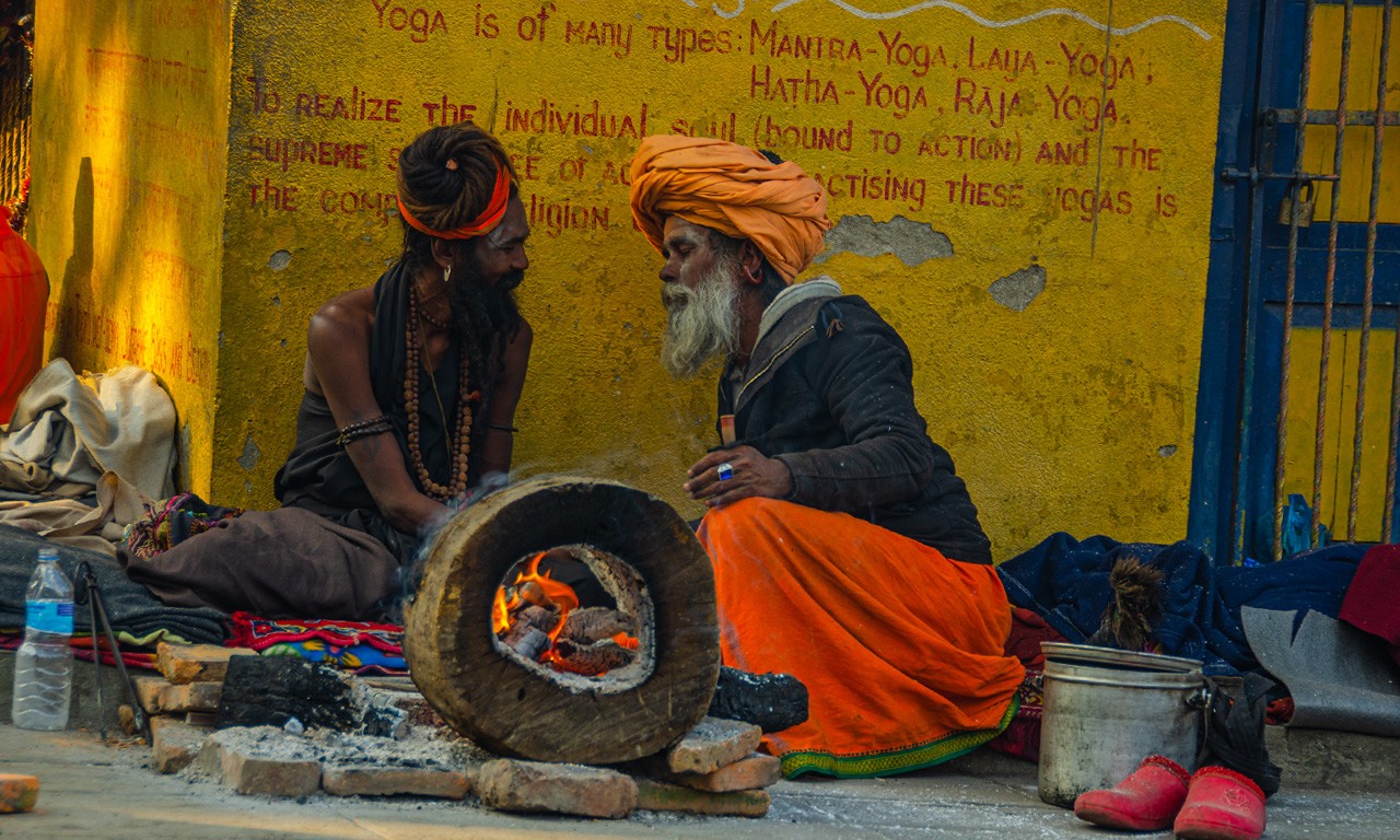 Two Sadhus with traditional turbans and beads sitting by a small ritual fire at Pashupatinath, symbolizing renunciation and devotion amidst a yellow wall with spiritual inscriptions.