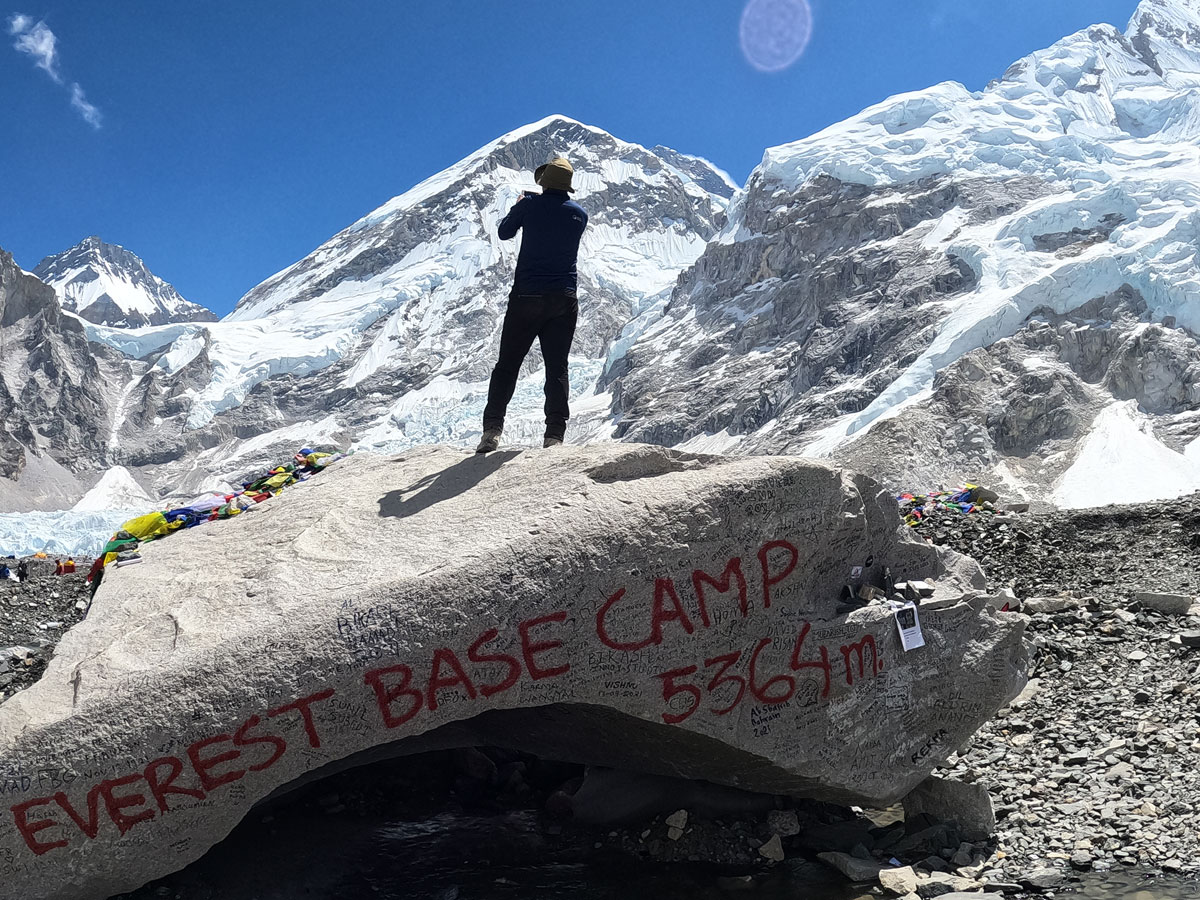 A trekker taking a photo standing on the iconic Everest Base Camp rock marker painted with EBC 5,364m on the Khumbu Glacier