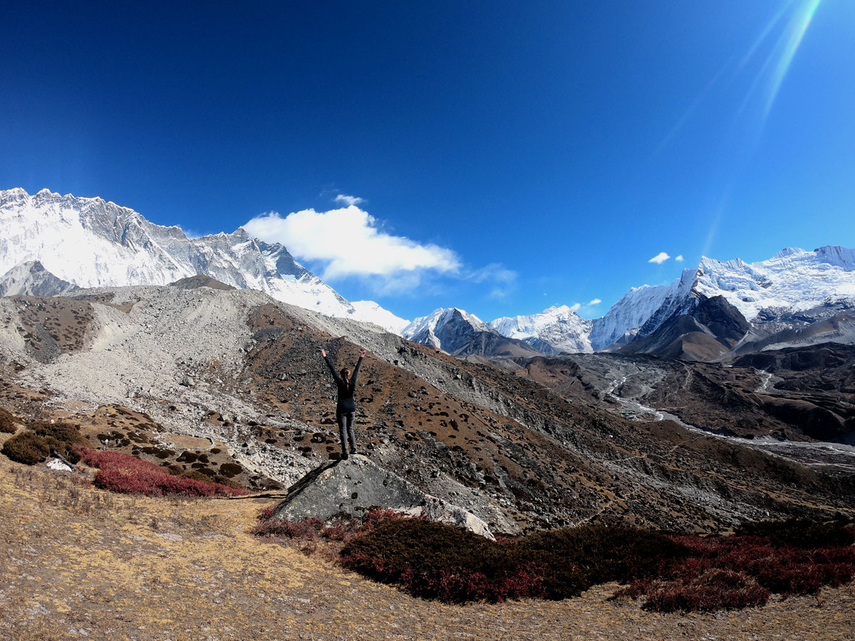 A hiker standing on a rock with arms outstretched celebrating surrounded by vast alpine slopes and snow-draped peaks