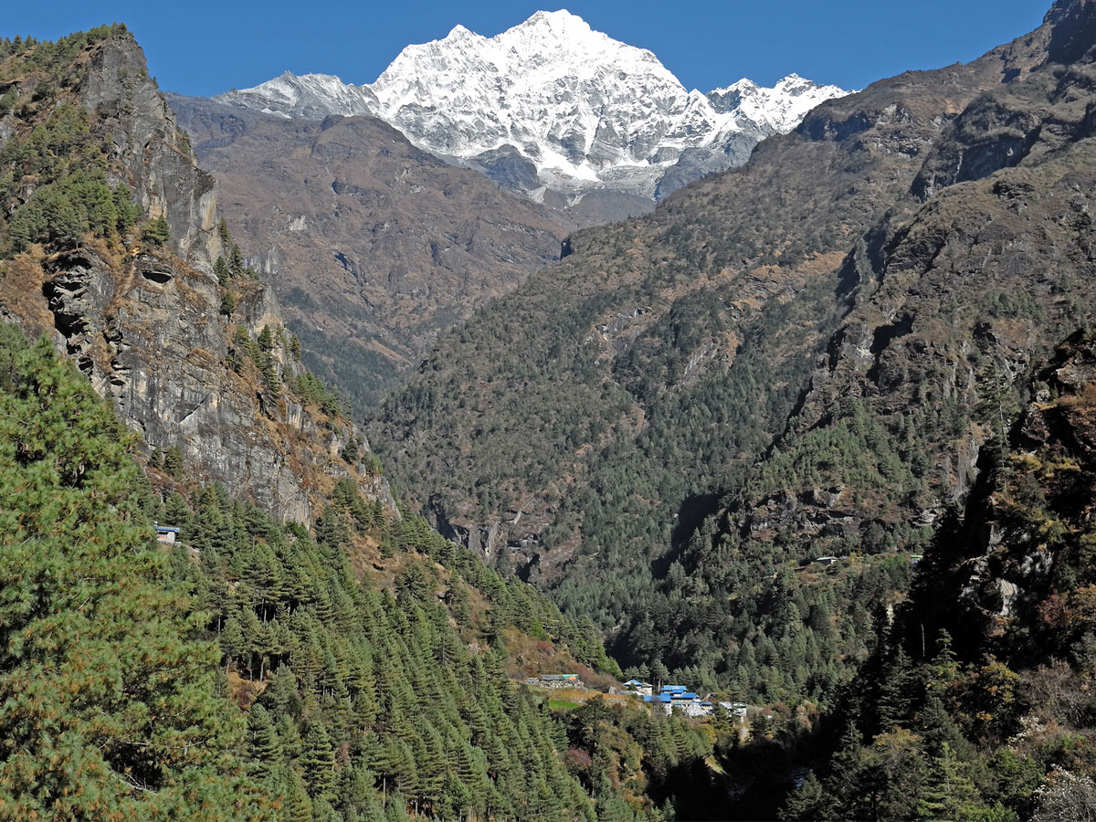 Dense green pine forest canyon with small traditional village roofs in the gorge and the towering snow-capped peak of Thamserku rising in the background