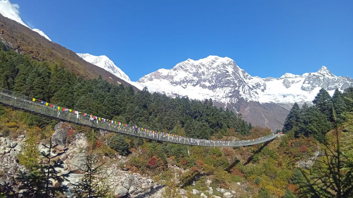Suspension bridge decorated with prayer flags on the Manaslu Circuit trek between Shyala and Samagaon under clear blue skies.