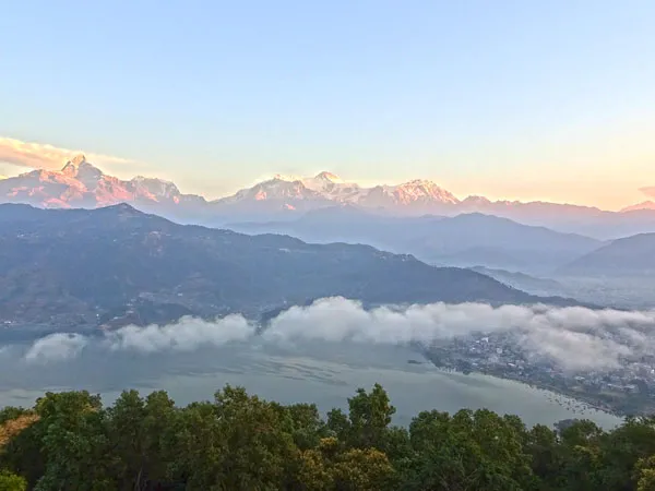 A high-angle panoramic sunrise view overlooking Phewa Lake with low-lying morning clouds, Pokhara city, and the snow-capped Annapurna mountain range glowing in soft morning light