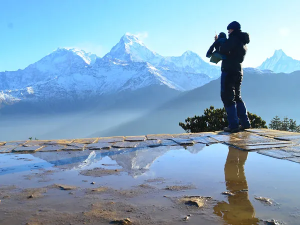 A hiker standing on a stone paving terrace looking towards snow-capped Himalayan peaks, with their reflection and mountain reflection visible in a clear water puddle on the ground