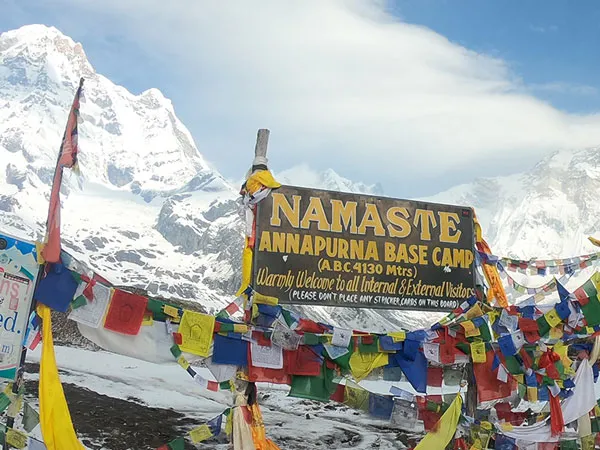 Welcome signboard at Annapurna Base Camp located at 4,130 meters elevation, surrounded by colorful Buddhist prayer flags and snowy Himalayan peaks in Nepal