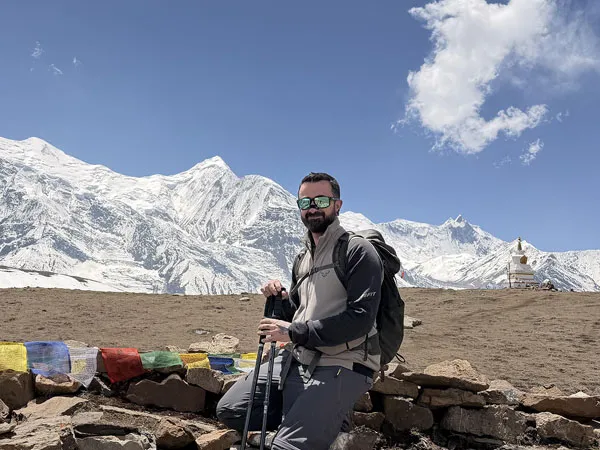 Trekker with trekking poles and backpack sitting near Buddhist prayer flags with snow-capped Annapurna mountain peaks and a stupa in Nepal