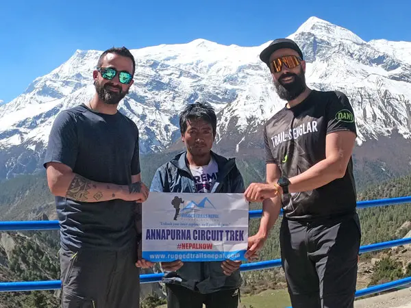 Three men holding an Annapurna Circuit Trek banner with snow-capped mountains in the background.