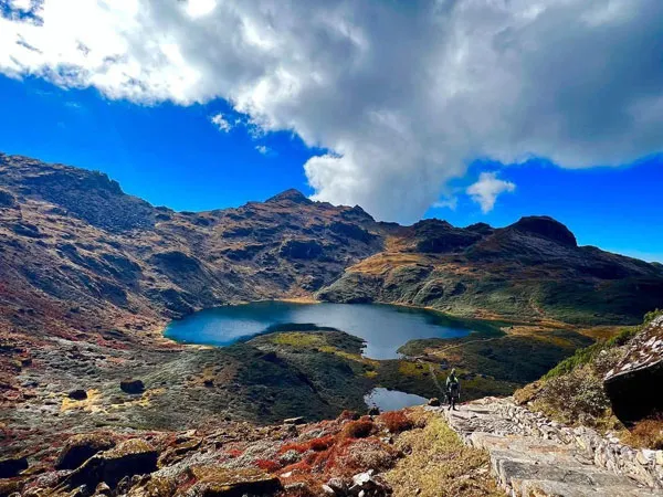 A high-angle view of a deep blue glacial alpine lake nestled in a rocky valley beneath mountain peaks, with a stone-paved hiking trail leading through the foreground under a dynamic blue sky with fluffy white clouds