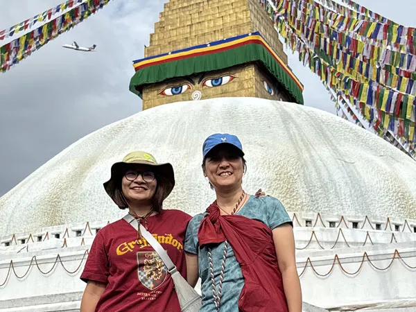 Two female tourists smiling in front of the white dome and eye-painted spire of Boudhanath Stupa, decorated with colorful prayer flags stretching across the sky