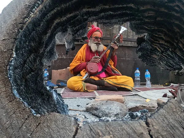 A Hindu Sadhu holy man in saffron robes, turban, and sunglasses sitting cross-legged and playing an ektara string instrument, framed naturally through the hollow opening of a charred tree trunk near Pashupatinath Temple