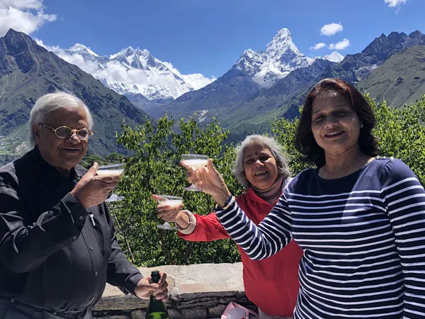 Three travelers raising champagne glasses in a toast on an outdoor terrace, with the snow-covered peaks of Mount Everest, Lhotse, and Ama Dablam visible in the background
