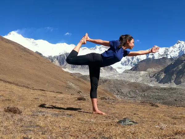 A trekker holding Natarajasana (Dancer Pose) outdoors with snow-covered Himalayan mountains behind her.