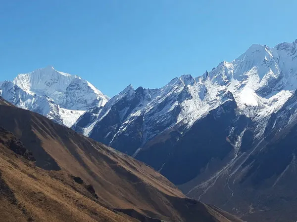 Panoramic view of rugged brown mountain slopes leading up to majestic, snow-covered Himalayan peaks in the Langtang region under a clear blue sky