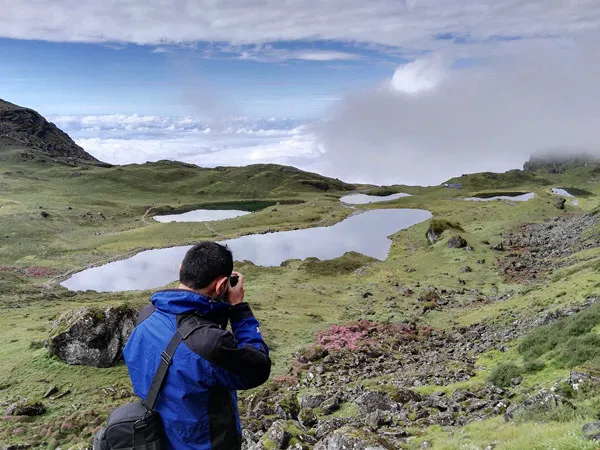 A trekker in a blue jacket taking a photo of the sacred alpine lakes of Panch Pokhari nestled among green hills in Nepal