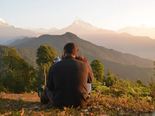 A couple sitting together on a grassy hill from behind, watching the golden morning sunlight illuminate the peak of Machhapuchhre (Fishtail) mountain and the Annapurna range